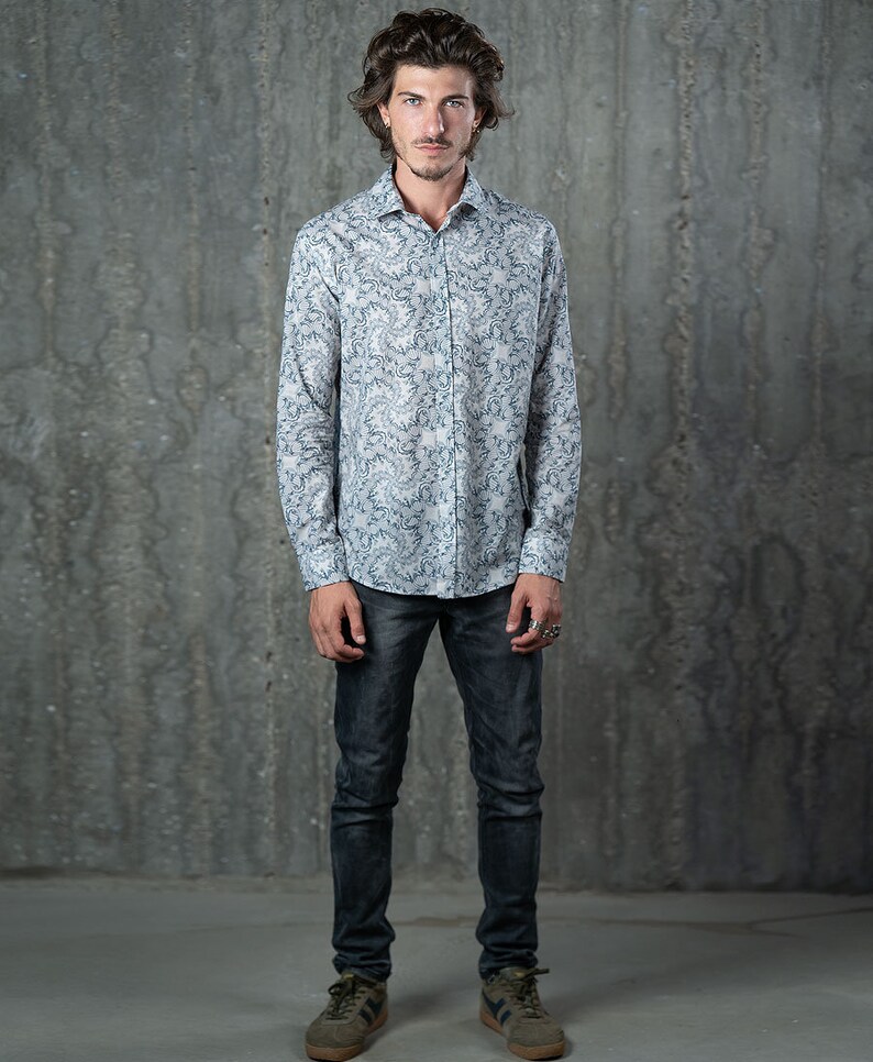 May include: A man wearing a white long-sleeved shirt with a blue and white floral pattern. He is wearing dark blue jeans and green sneakers.