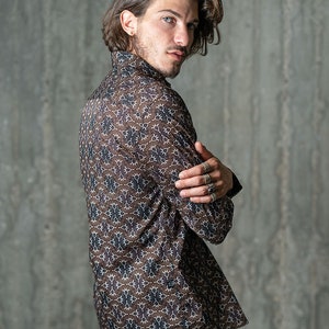 May include: A brown button-down shirt with a repeating floral pattern in shades of white and navy blue. The long-sleeved shirt is worn with the arms crossed. The shirt is made of a lightweight material.