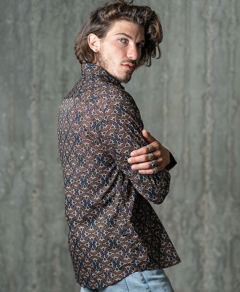 May include: A brown button-down shirt with a repeating floral pattern in shades of white and navy blue. The long-sleeved shirt is worn with the arms crossed. The shirt is made of a lightweight material.