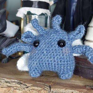 May include: A handmade blue crochet creature with black button eyes and white accents. The whimsical amigurumi figure has multiple arms and is displayed on a wooden surface. The toy is a unique, handcrafted item.