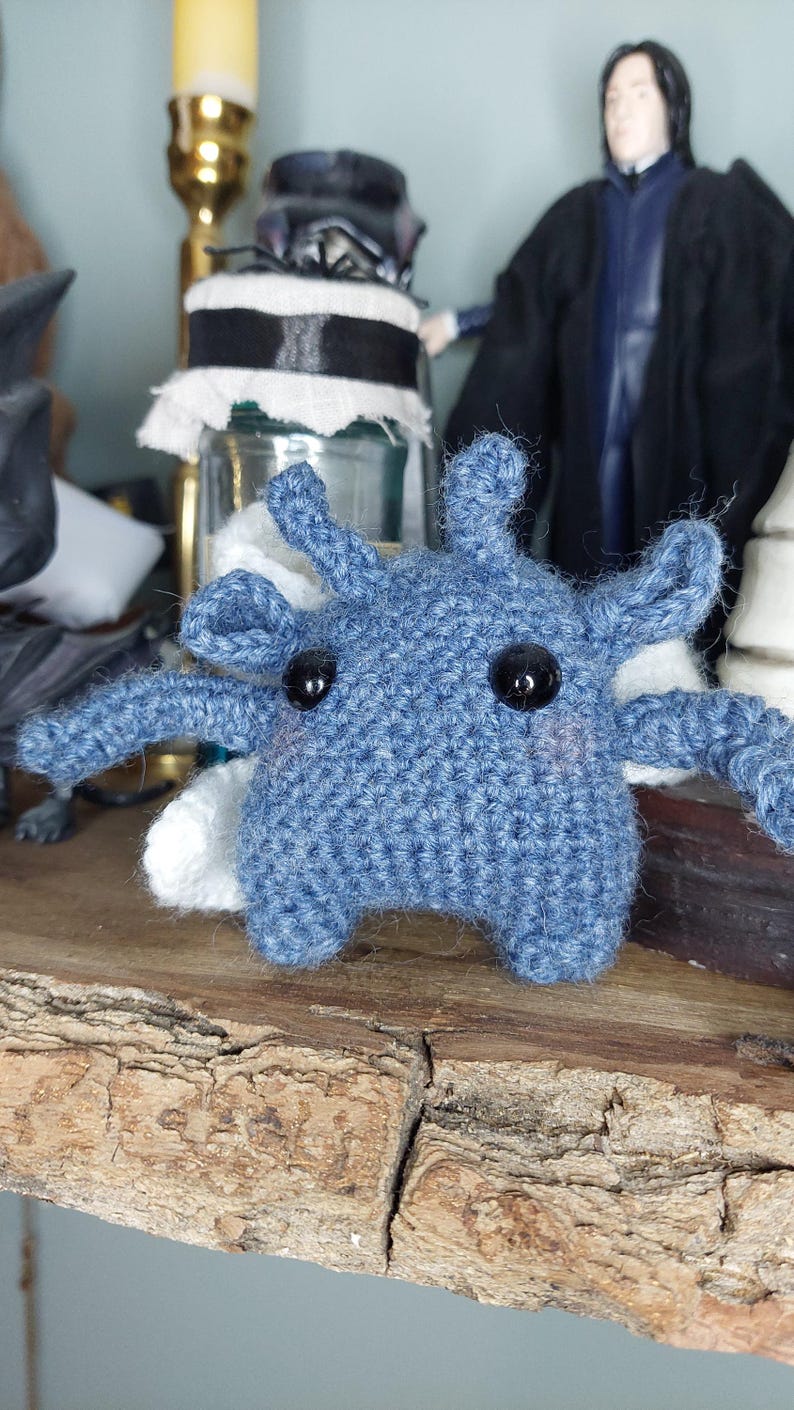 May include: A handmade blue crochet creature with black button eyes and white accents. The whimsical amigurumi figure has multiple arms and is displayed on a wooden surface. The toy is a unique, handcrafted item.