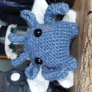 May include: A handmade blue crochet octopus with black button eyes. The octopus is attached to a white and black object. The background includes a wooden surface and other decorative items.