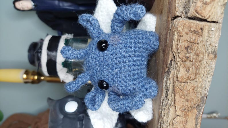 May include: A handmade blue crochet octopus with black button eyes. The octopus is attached to a white and black object. The background includes a wooden surface and other decorative items.