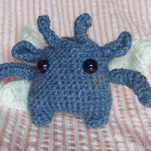 May include: A handmade blue crochet creature with black button eyes and white wings. The plush toy has four arms and four antennae. It is resting on a pink and white striped fabric.