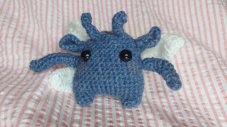 May include: A handmade blue crochet creature with black button eyes and white wings. The plush toy has four arms and four antennae. It is resting on a pink and white striped fabric.