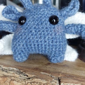May include: A handmade, blue crochet creature with black bead eyes and white accents. The plush toy has four appendages and two antennae. It is displayed on a wooden surface.
