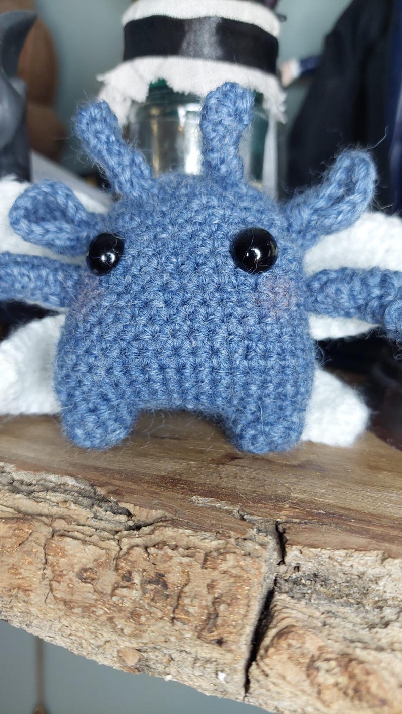 May include: A handmade, blue crochet creature with black bead eyes and white accents. The plush toy has four appendages and two antennae. It is displayed on a wooden surface.