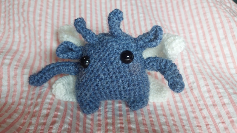 May include: A handmade blue crochet creature with black button eyes and white wings. The plush toy has multiple appendages and is set against a pink and white striped background. The toy is a unique, whimsical creation.