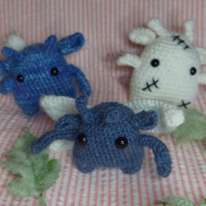 May include: Three handmade crochet creatures in blue and white, with black button eyes and stitched details. The creatures are arranged on a pink and white striped background, with a purple flower and green leaf accents.