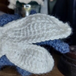 May include: Close-up of a pair of handmade crochet mittens. The mittens feature a white, textured design on the top and a blue base. The mittens are made of soft yarn and appear warm and cozy.