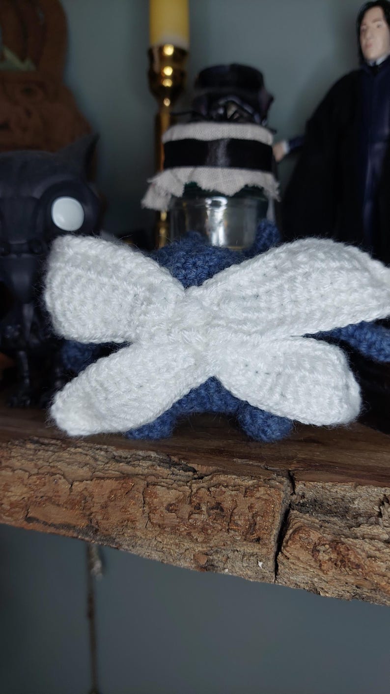 May include: A handmade crochet dragonfly with white wings and a blue body. The wings are large and crossed, and the dragonfly is sitting on a wooden surface. This whimsical item is perfect for home decor or as a unique gift.