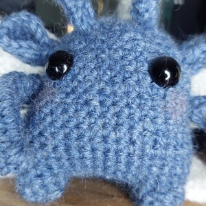 May include: A close-up of a handmade blue crochet creature with black button eyes. The plush toy has a textured surface and small, pointed ears. The background is blurred, with hints of other objects.