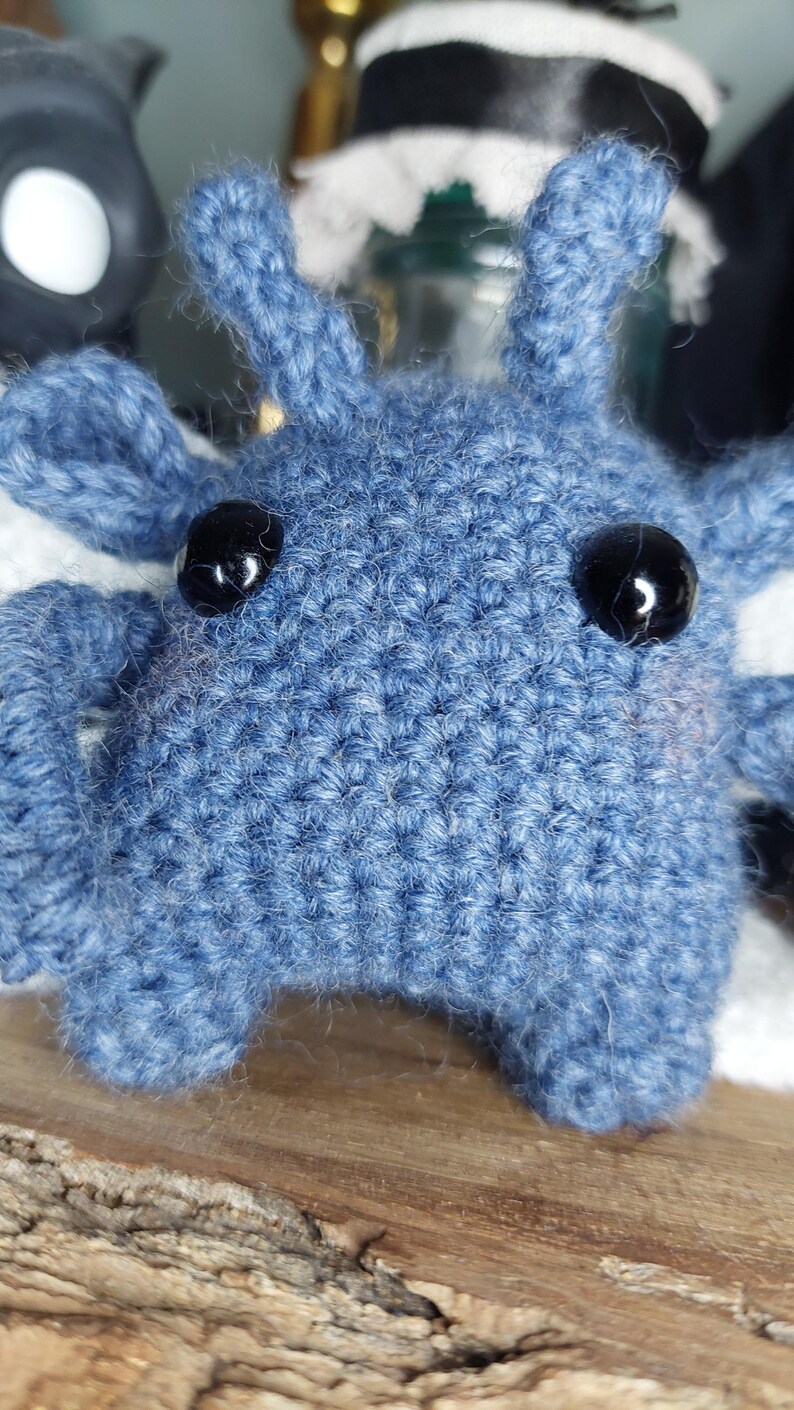 May include: A close-up of a handmade blue crochet creature with black button eyes. The plush toy has a textured surface and small, pointed ears. The background is blurred, with hints of other objects.