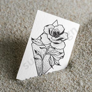 May include: Black and white rose illustration on a white paper. The rose is inside a geometric shape with a stem and leaves. The paper is partially buried in a bed of small, light-colored pebbles.