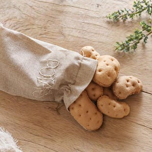May include: A beige linen bag with the word "POTATO" embroidered on it, spilling out several tan-colored felt potatoes. The potatoes have small brown stitched details. The scene is set on a light brown wooden surface.