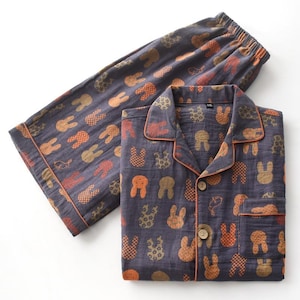 K&ouml;nnte beinhalten: Ein Set grauer Pyjamas mit einem sich wiederholenden Muster aus orangefarbenen und braunen Hasensilhouetten. Das Hemd mit Kn&ouml;pfen und die Hose haben einen orangefarbenen Besatz. Die Pyjamas sind aus einem weichen, leichten Stoff.