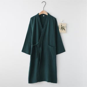 Può includere: Un accappatoio verde scuro con scollo a V e maniche lunghe è appeso a un'appendiabiti di legno. L'accappatoio ha una cintura annodata e due tasche anteriori. Il tessuto sembra essere una miscela di cotone o lino strutturato. L'accappatoio è esposto contro un muro bianco.