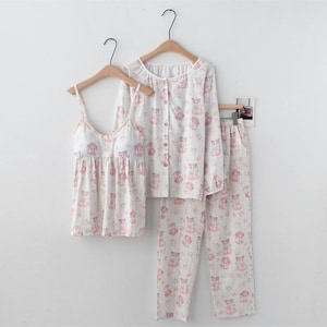 May include: A three-piece pajama set in white with a repeating pink bear pattern. The set includes a camisole, a button-down long-sleeve shirt, and matching trousers. The camisole has spaghetti straps and a lace trim. The pajamas are hanging on wooden hangers.