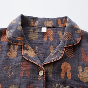 K&ouml;nnte beinhalten: Nahaufnahme eines dunkelgrauen Pyjama-Oberteils mit einem sich wiederholenden Muster aus braunen, orangefarbenen und gr&uuml;nen Hasen. Der Kragen und die Kanten sind mit einer orangefarbenen Paspel versehen. Das Oberteil hat eine Knopfleiste und eine einzelne Tasche.