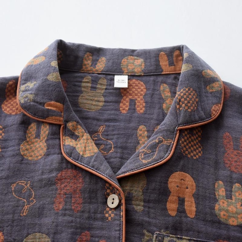 K&ouml;nnte beinhalten: Nahaufnahme eines dunkelgrauen Pyjama-Oberteils mit einem sich wiederholenden Muster aus braunen, orangefarbenen und gr&uuml;nen Hasen. Der Kragen und die Kanten sind mit einer orangefarbenen Paspel versehen. Das Oberteil hat eine Knopfleiste und eine einzelne Tasche.