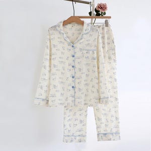 Könnte beinhalten: Cremefarbenes Pyjama-Set mit einem sich wiederholenden blauen Katzenmuster. Das langärmlige Oberteil hat blaue Knöpfe und Besätze, und die Hose hat einen passenden Besatz. Das Set hängt an einem Holzbügel.