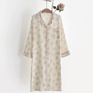 Pode incluir: Um roupão creme de manga comprida com um padrão floral em rosa e verde. O roupão tem gola, um bolso frontal e acabamentos rosa nas bordas. Está pendurado num cabide de madeira.