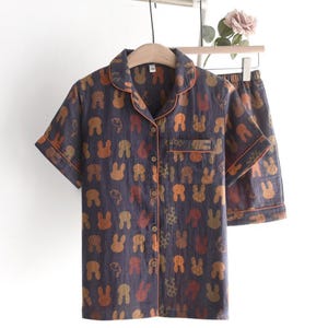 K&ouml;nnte beinhalten: Marineblaues Pyjama-Set mit einem sich wiederholenden Muster aus orangefarbenen und braunen Hasengesichtern. Das kurz&auml;rmelige Button-Down-Hemd und die passenden Shorts haben einen orangefarbenen Besatz. Das Set h&auml;ngt an einem Holzb&uuml;gel.