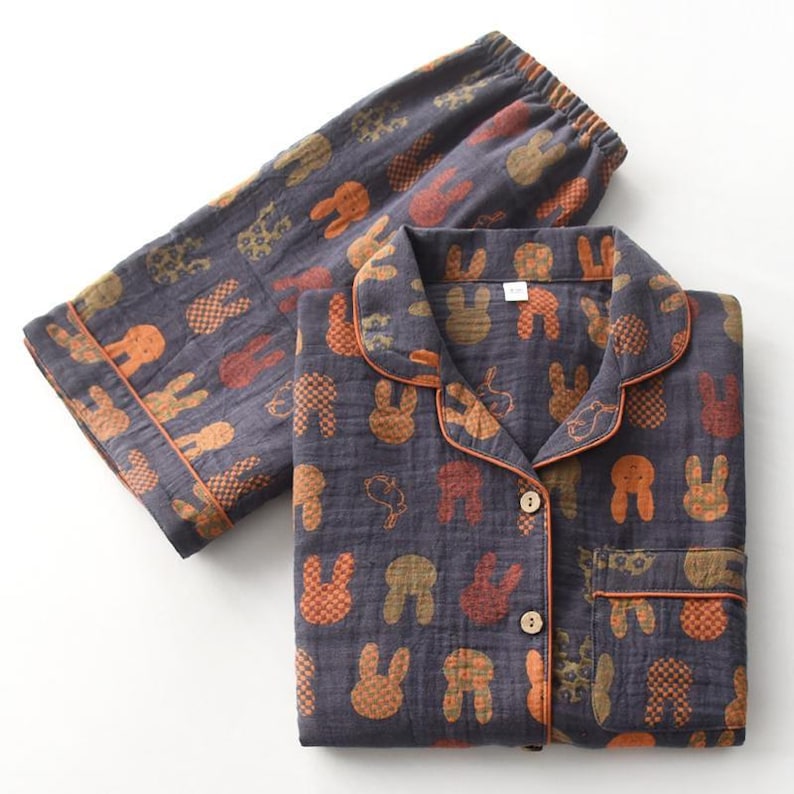 K&ouml;nnte beinhalten: Dunkelgraues Pyjama-Set mit einem sich wiederholenden Muster aus orangefarbenen, braunen und beigen Hasensilhouetten. Das Set beinhaltet ein Hemd mit Kn&ouml;pfen und orangefarbenem Besatz sowie eine passende Hose mit elastischem Bund.