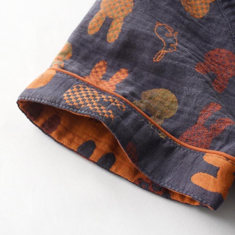 K&ouml;nnte beinhalten: Nahaufnahme eines Pyjama-&Auml;rmels. Der &Auml;rmel ist dunkelgrau mit einem Muster aus orangefarbenen und gr&uuml;nen Hasen und V&ouml;geln. Das B&uuml;ndchen ist orange mit einem geometrischen Muster und braunem Besatz.