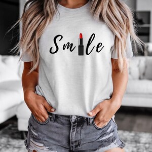May include: A light gray t-shirt with the word "Smile" in black script, featuring a red lipstick graphic. The shirt is a casual style, suitable for everyday wear.