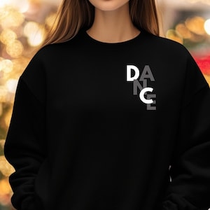 Puede incluir: Sudadera negra con la palabra "DANCE" impresa en blanco y gris. Las letras están apiladas verticalmente, con "D" y "C" en blanco y "A", "N" y "E" en gris. La sudadera es de color negro liso.