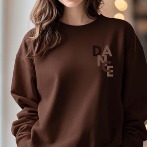 Puede incluir: Una sudadera marrón con la palabra "DANCE" impresa verticalmente en un degradado de tonos marrones en la parte superior izquierda del pecho. La sudadera tiene mangas largas y cuello redondo.