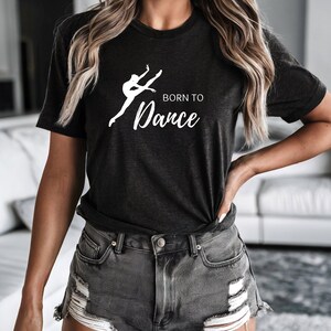 Può includere: Una t-shirt a maniche corte color grigio antracite con una grafica bianca che raffigura una persona che danza in posa di salto. Il testo "BORN TO" è stampato in un carattere sans-serif sopra la parola "Dance" scritta in corsivo bianco.