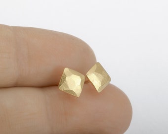 May include: A pair of gold-colored, square stud earrings. Each earring has a textured, faceted surface, catching the light. The earrings are displayed against a neutral background, highlighting their geometric design and metallic finish.
