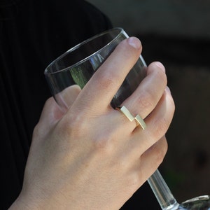 May include: A gold ring with a simple, geometric design. The ring is made of two rectangular pieces of metal that are joined together at a right angle.