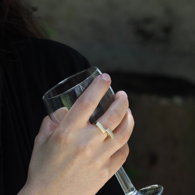 May include: A gold ring with a simple, geometric design. The ring is made of two rectangular pieces of metal that are joined together at a right angle.