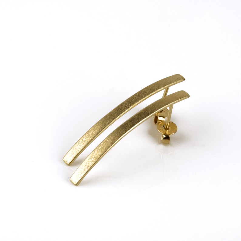 May include: A pair of gold-toned earrings with a curved, brushed finish. The earrings are made of two thin, parallel bars.
