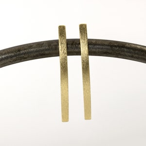 May include: A pair of gold-toned earrings with a brushed finish. The earrings are long and thin, and they are hanging from a black stand.