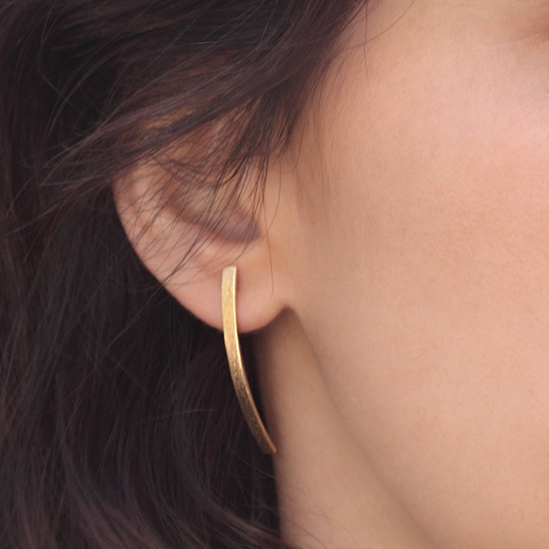 May include: A pair of gold-toned earrings with a long, thin, curved design. The earrings are worn in the earlobes.