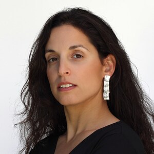 May include: A woman with long dark hair wearing silver dangle earrings. The earrings are made of multiple stacked squares.