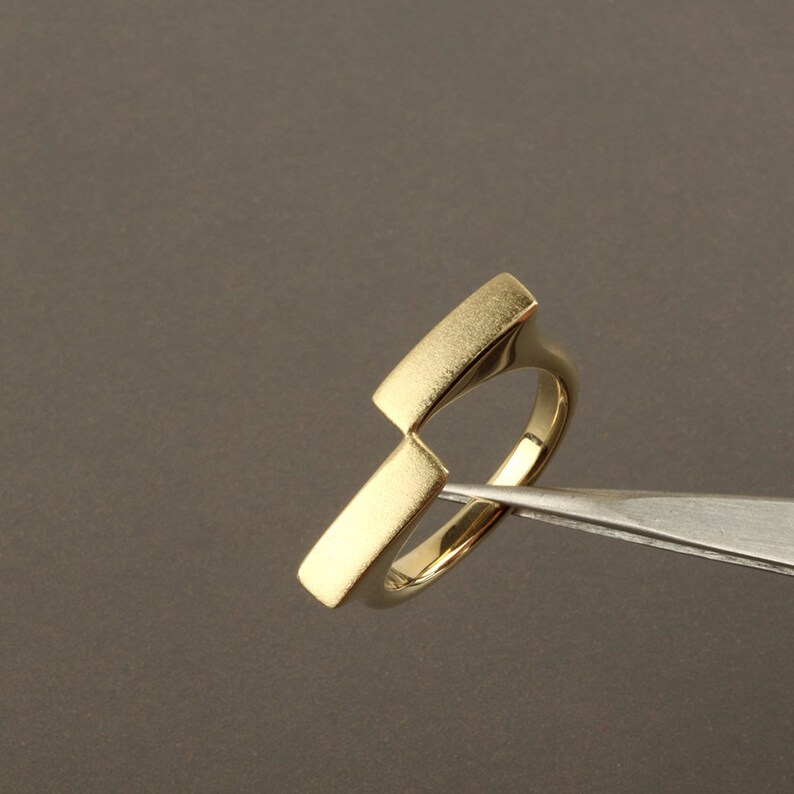 May include: A gold ring with a unique, geometric design. The ring is made of two rectangular pieces of metal that are joined together at an angle.