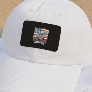 May include: A white baseball cap with a curved brim, featuring a black rectangular patch. The patch displays an eagle, American flag, and the text "1776-2020". The cap is resting on a white t-shirt, all on a sandy surface.