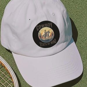 May include: White baseball cap with a circular patch featuring a cowboy on horseback, the text "COWBOY STATE OF MIND", a tennis racket, and a tennis ball on a green surface.