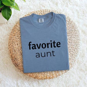 May include: A blue t-shirt with the words "favorite aunt" printed in black. The shirt is displayed on a round, woven mat, with a potted plant in the background. The shirt is a crew neck style.