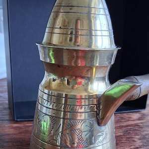 May include: A vintage brass coffee pot with a conical lid and a long wooden handle. The pot features etched floral patterns around the base and horizontal bands. The pot is designed for brewing coffee.