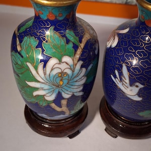May include: Two small cloisonné vases featuring a deep blue background with gold wire detailing. The vases display white and light blue floral patterns, green leaves, and a white bird motif. Each vase is set on a circular dark brown wooden base.