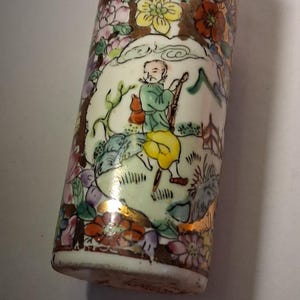 Pode incluir: Um recipiente cilíndrico de cerâmica com uma cena pintada à mão de uma figura com túnica verde e calças amarelas caminhando por uma paisagem. O design inclui motivos florais coloridos, detalhes dourados e uma borda decorativa superior.