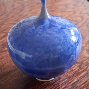 May include: A round ceramic vase with a narrow neck, featuring a glossy cobalt blue glaze with crystalline patterns that resemble starbursts or flowers. The base of the vase is white, and the surface has a smooth, reflective finish throughout.