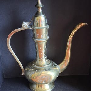 Peut inclure: Une cruche en laiton vintage au design orné. La cruche présente un long bec incurvé, une poignée et un couvercle décoratif. Le laiton a une teinte dorée chaude et la surface est gravée de motifs complexes. La cruche est probablement utilisée pour servir des boissons.