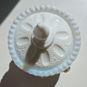 Puede incluir: Un portaanillos de vidrio blanco opalino con un cono central y una base redonda. La base presenta un patrón floral detallado y un borde con cuentas. El portaanillos está diseñado para guardar anillos y otras joyas pequeñas.